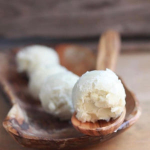 a wooden spoon rests on top of a rustic wooden bowl. The spoon and bowl each contains perfectly round balls of rich and moisturizing shea butter